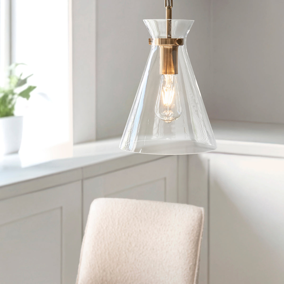 A modern pendant light with a clear glass shade and brass accents hangs above a light-colored chair.