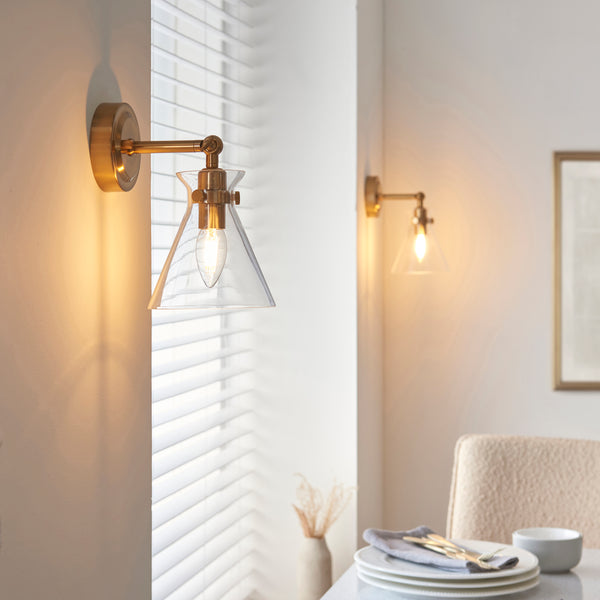 Two brass wall sconces with clear glass shades illuminate a dining area with a set table.