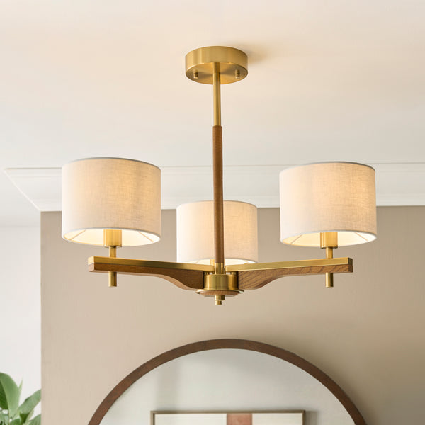 A brass and wood chandelier with three fabric shades illuminates a room with a mirror and plant.