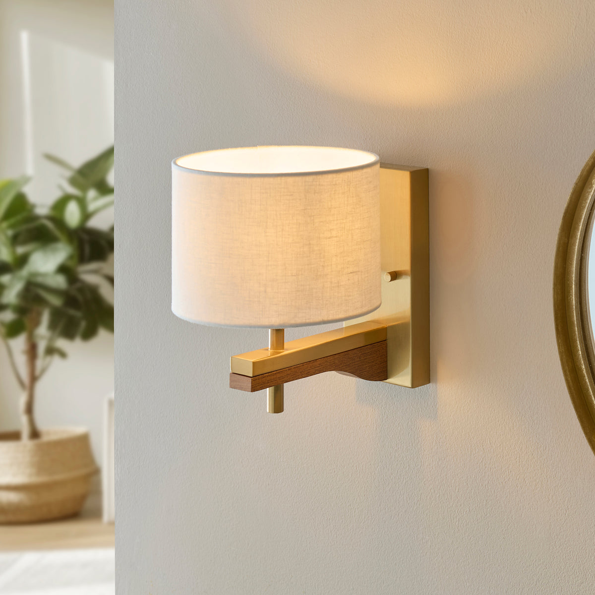 A modern wall sconce with a white fabric shade, brass accents, and a wooden arm, illuminating a light-colored wall.