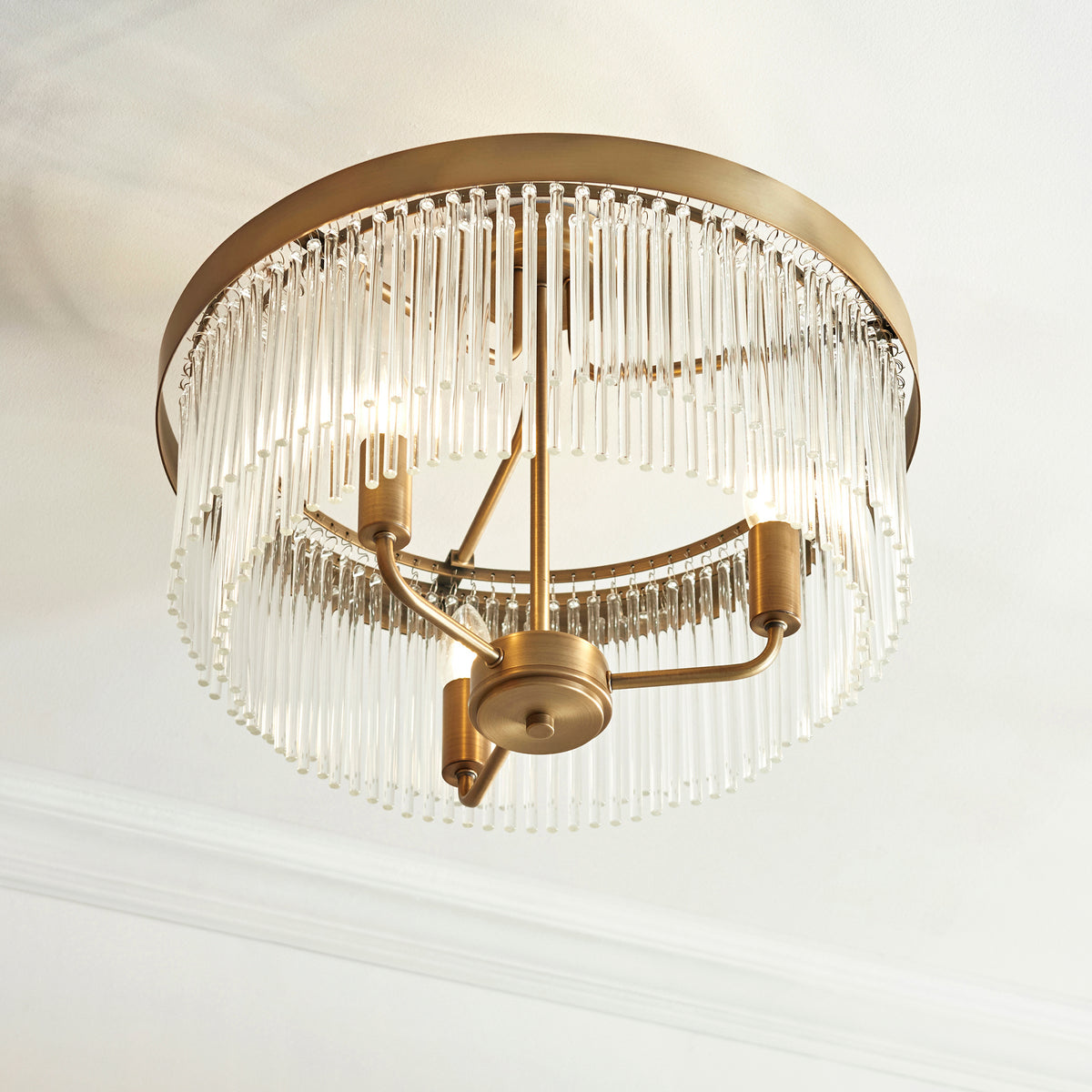 Round brass chandelier with clear glass rods hanging from the rim, illuminating a white ceiling.