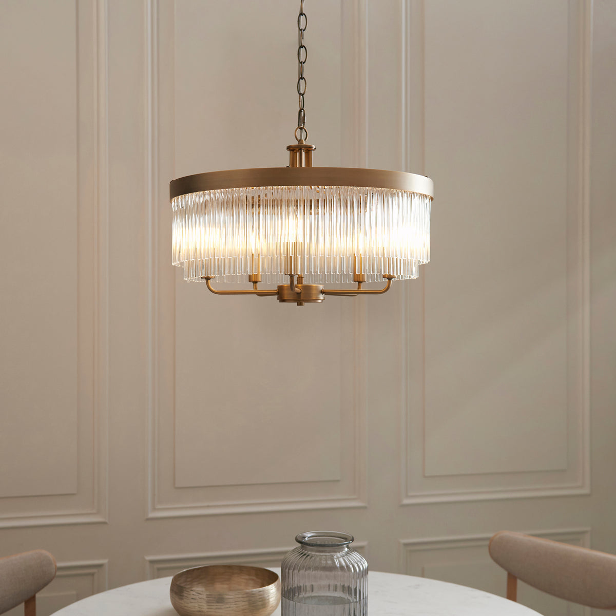 A modern brass chandelier with vertical glass rods hangs over a white marble table with a gold bowl and glass vase.