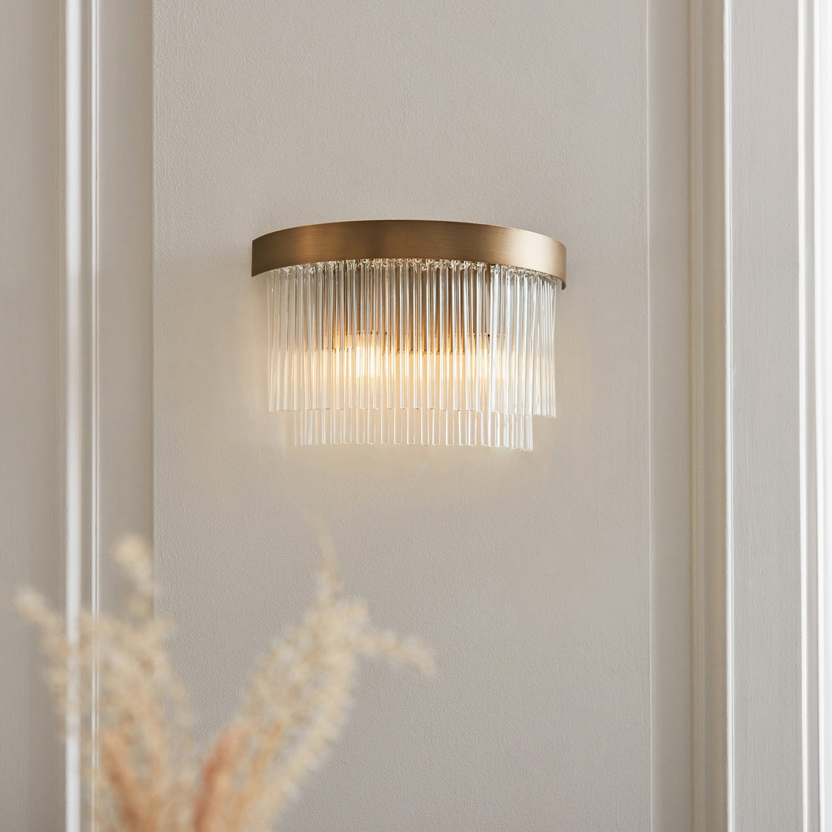 A modern brass wall sconce with clear fluted glass rods illuminates a light grey wall.