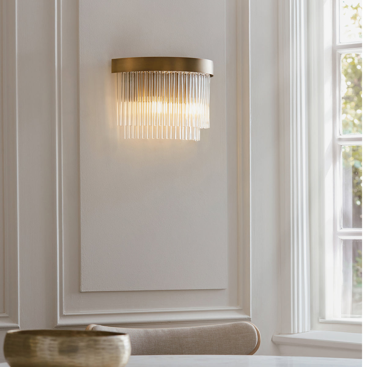 A brass wall sconce with clear glass rods illuminates a white paneled wall next to a window.