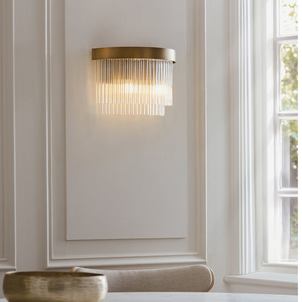 A brass wall sconce with clear glass rods illuminates a white paneled wall next to a window.