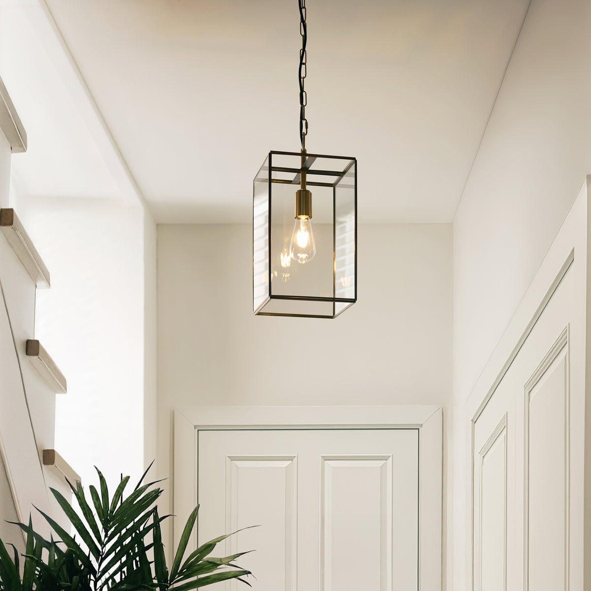A modern brass and glass lantern pendant light hangs in a bright hallway with white walls and doors.