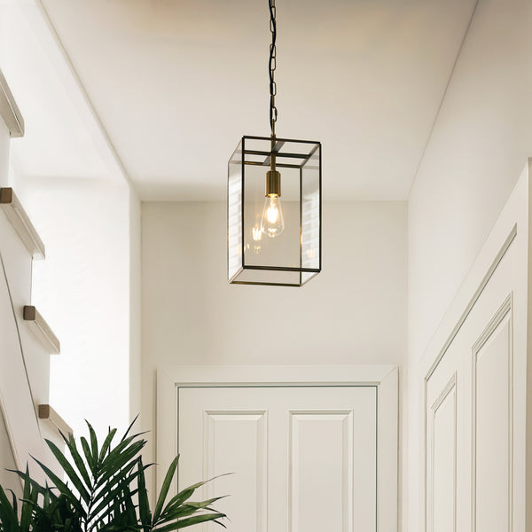 A modern brass and glass lantern pendant light hangs in a bright hallway with white walls and doors.