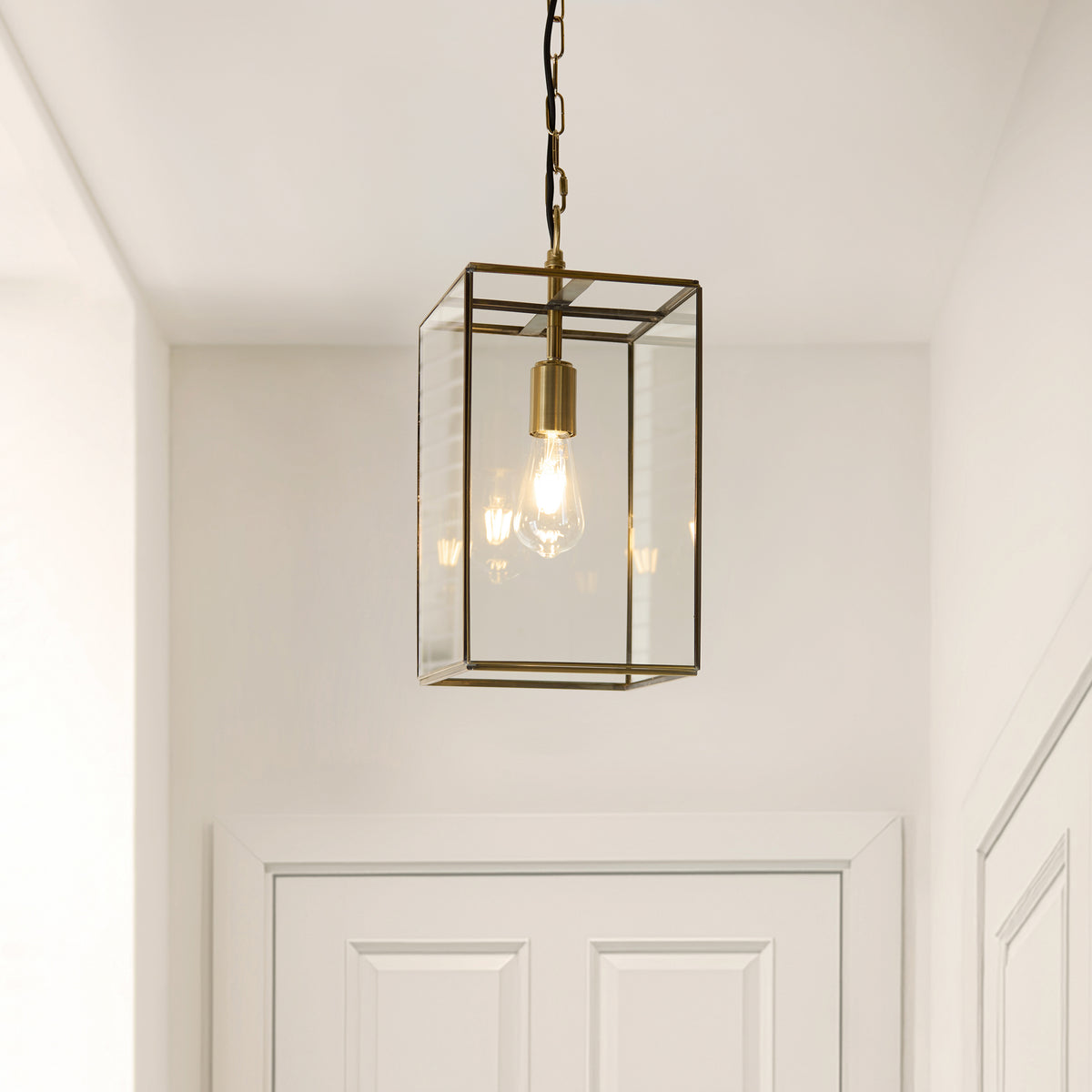 A brass and glass rectangular pendant light hangs from the ceiling, illuminating a white hallway with a white door.
