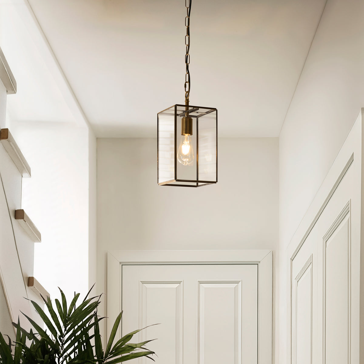 A modern brass and glass pendant light hangs in a bright hallway with white doors and a green plant.