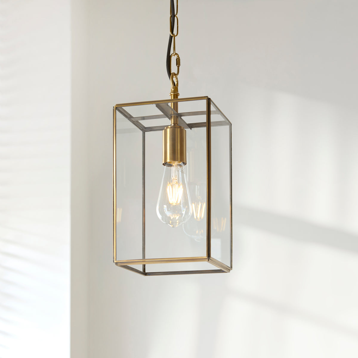 A brass and glass rectangular pendant light with an exposed filament bulb, hanging from a chain.