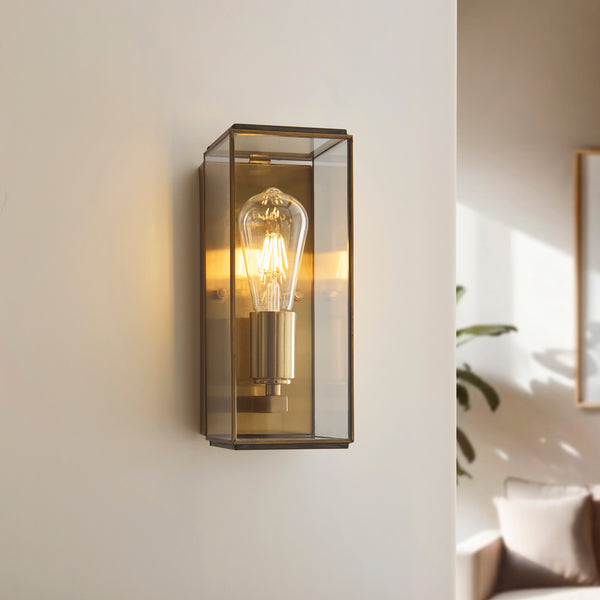 A brass and glass wall sconce with an exposed Edison bulb illuminates a light-colored wall.