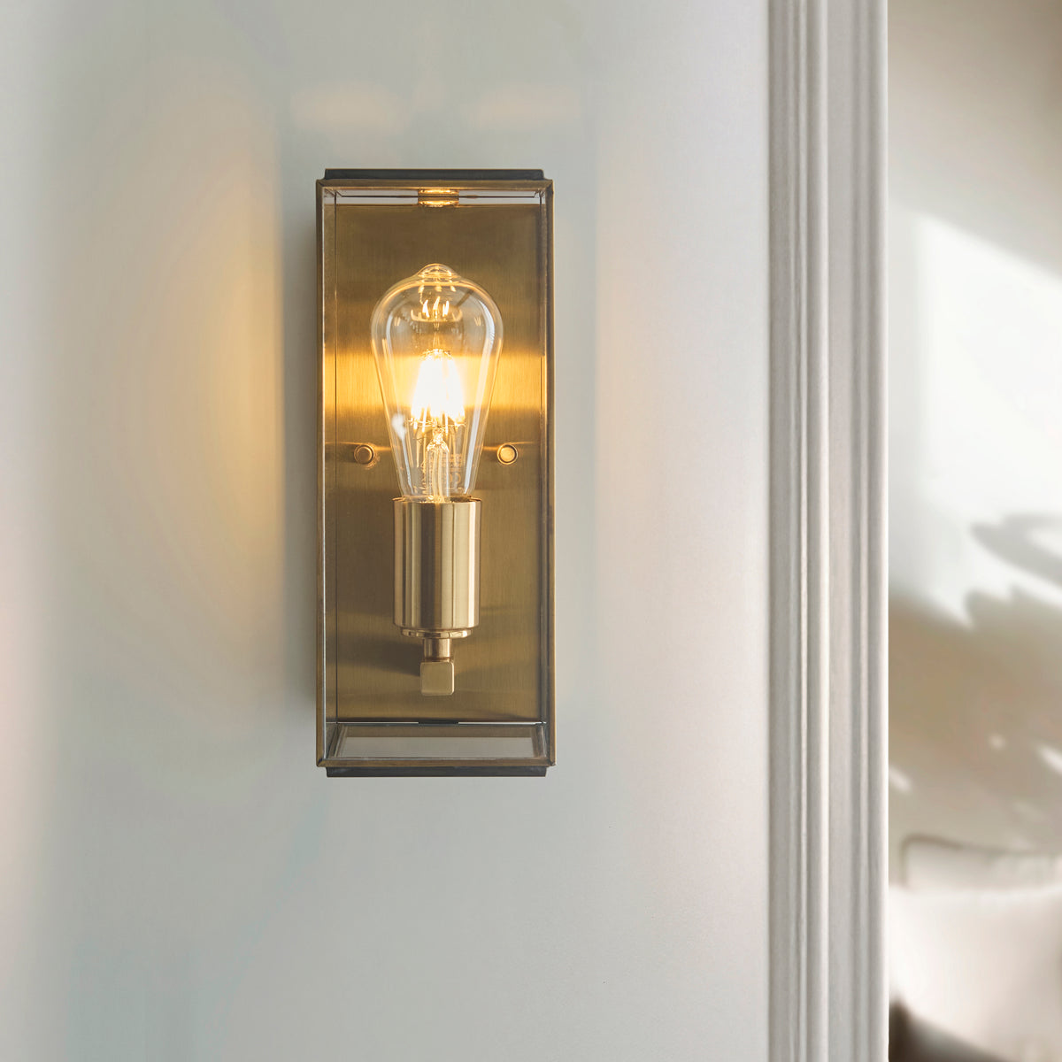 A brass wall sconce with a clear Edison bulb illuminates a light-colored wall, next to a white door frame.