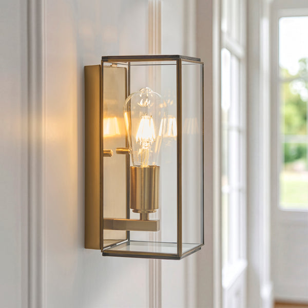 A brass and glass wall sconce with an exposed filament bulb, casting a warm glow on a white wall.