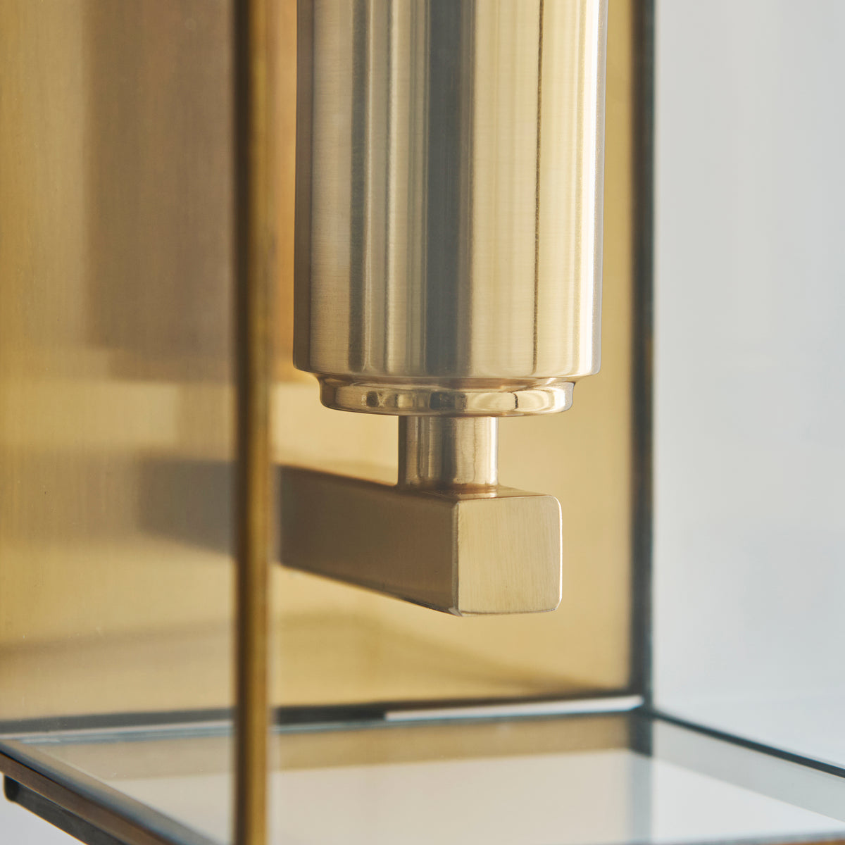 Close-up of a brass wall sconce with a cylindrical top and a rectangular arm, featuring glass panels.