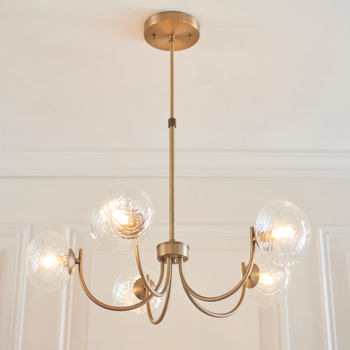 A gold chandelier with five clear, textured glass globes, each illuminated, hanging from a white ceiling.