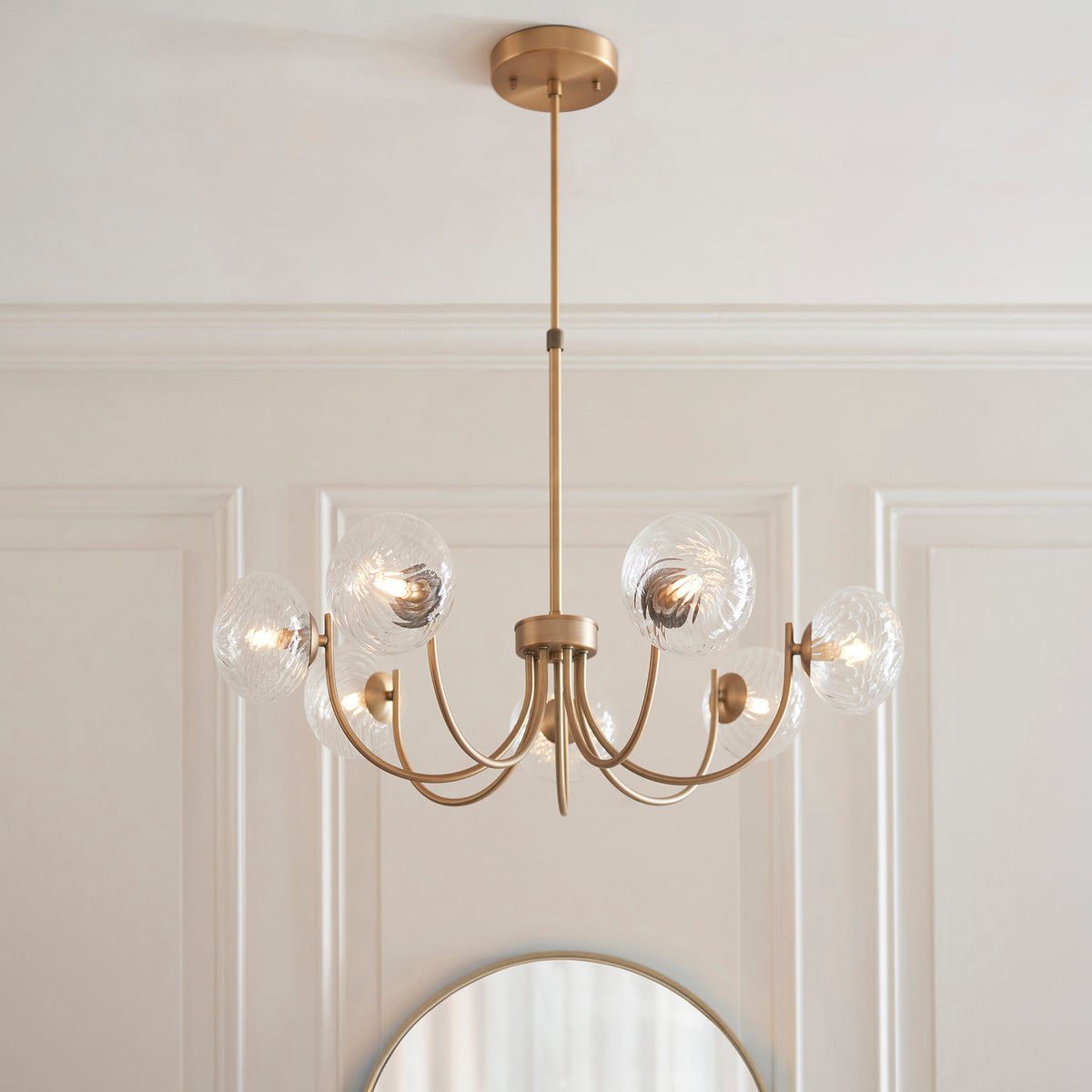 A gold chandelier with six textured glass globes hangs from a white ceiling above a round mirror.