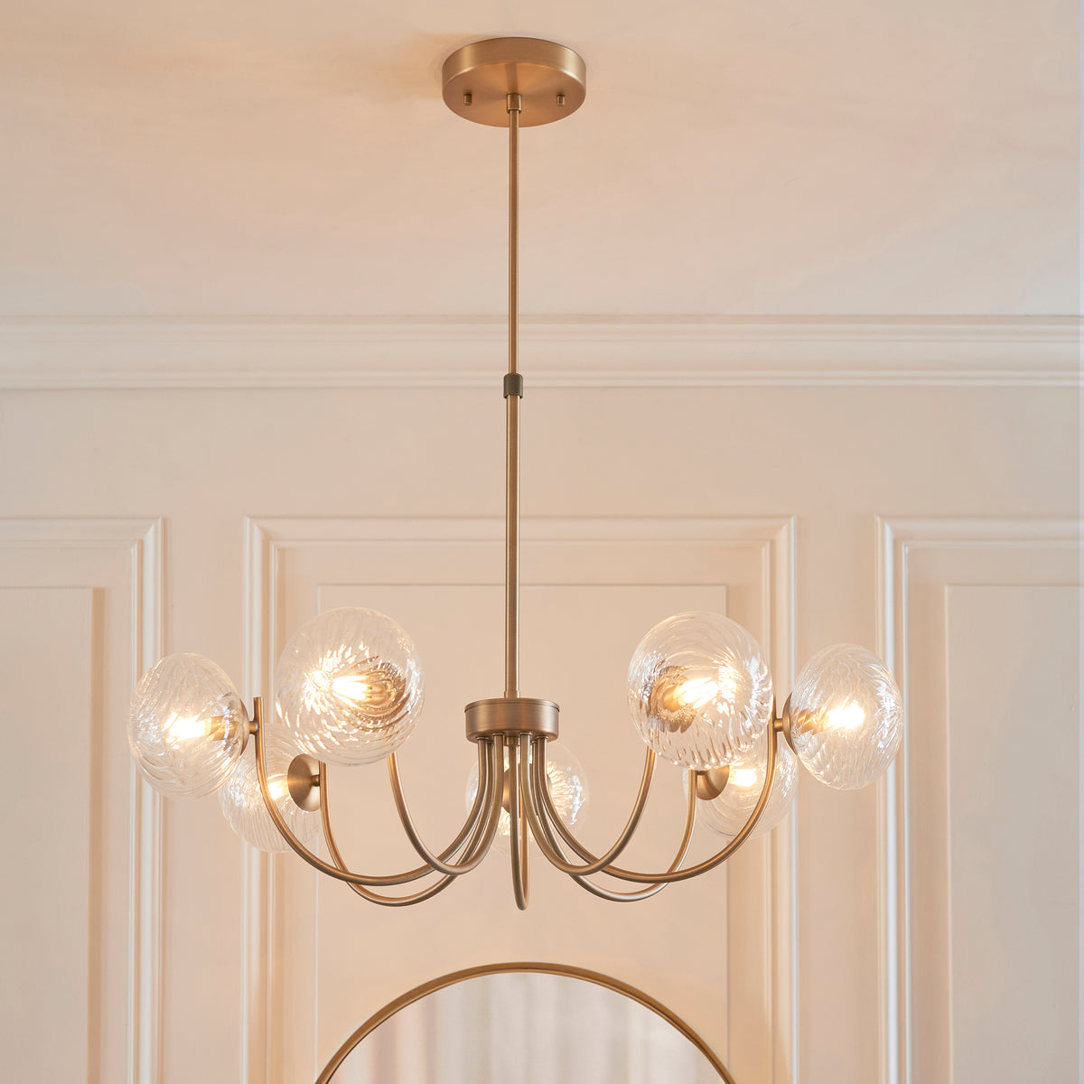 A gold chandelier with six clear, textured glass globes, each illuminated, hanging from a white ceiling.