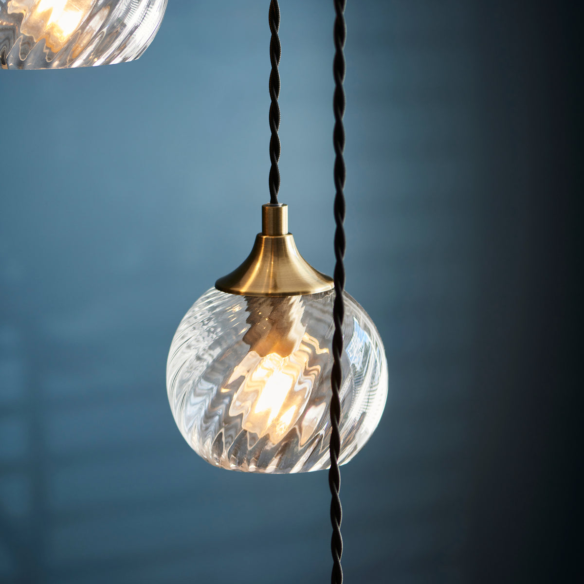 A close-up of a clear, textured glass pendant light with a brass fitting, glowing warmly against a blue wall.