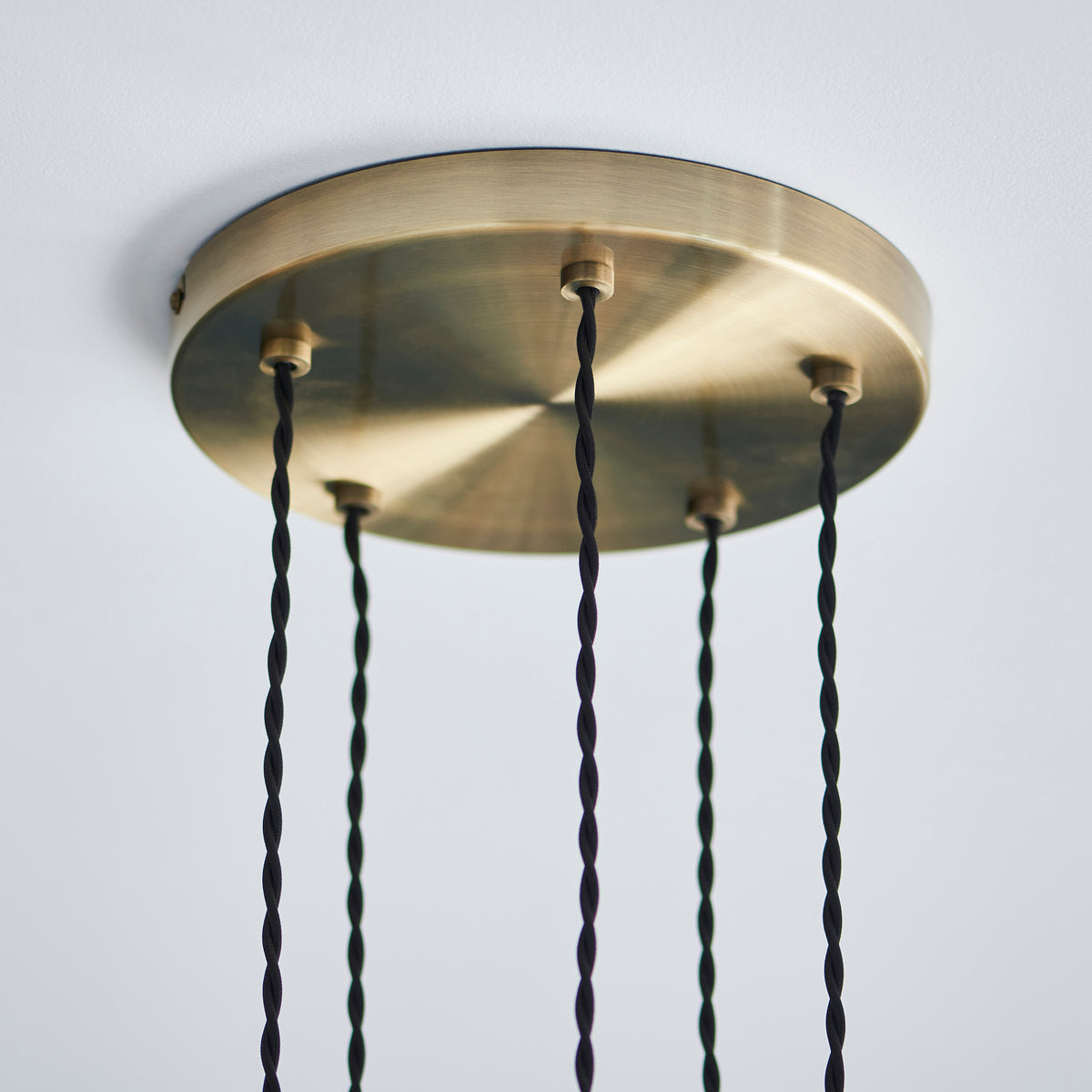 A close-up of a round, brushed brass ceiling rose with four black braided cables hanging down.