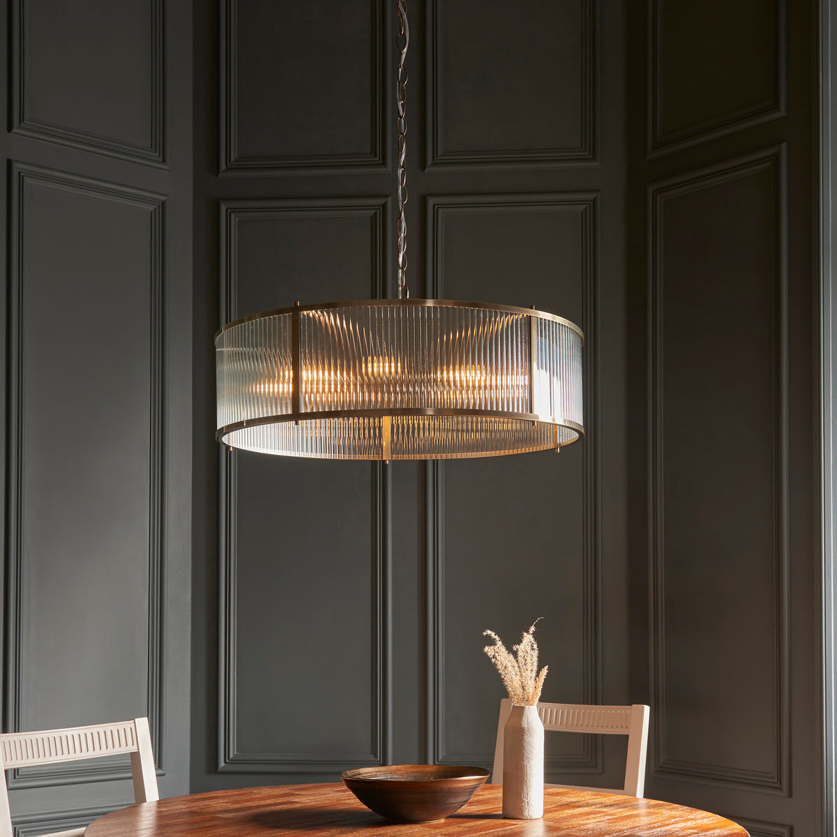 A dining room with dark paneled walls, a round wooden table, and a fluted glass chandelier.