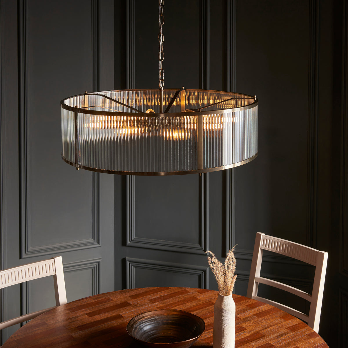 A round, fluted glass chandelier hangs above a wooden dining table with two chairs and a decorative bowl.