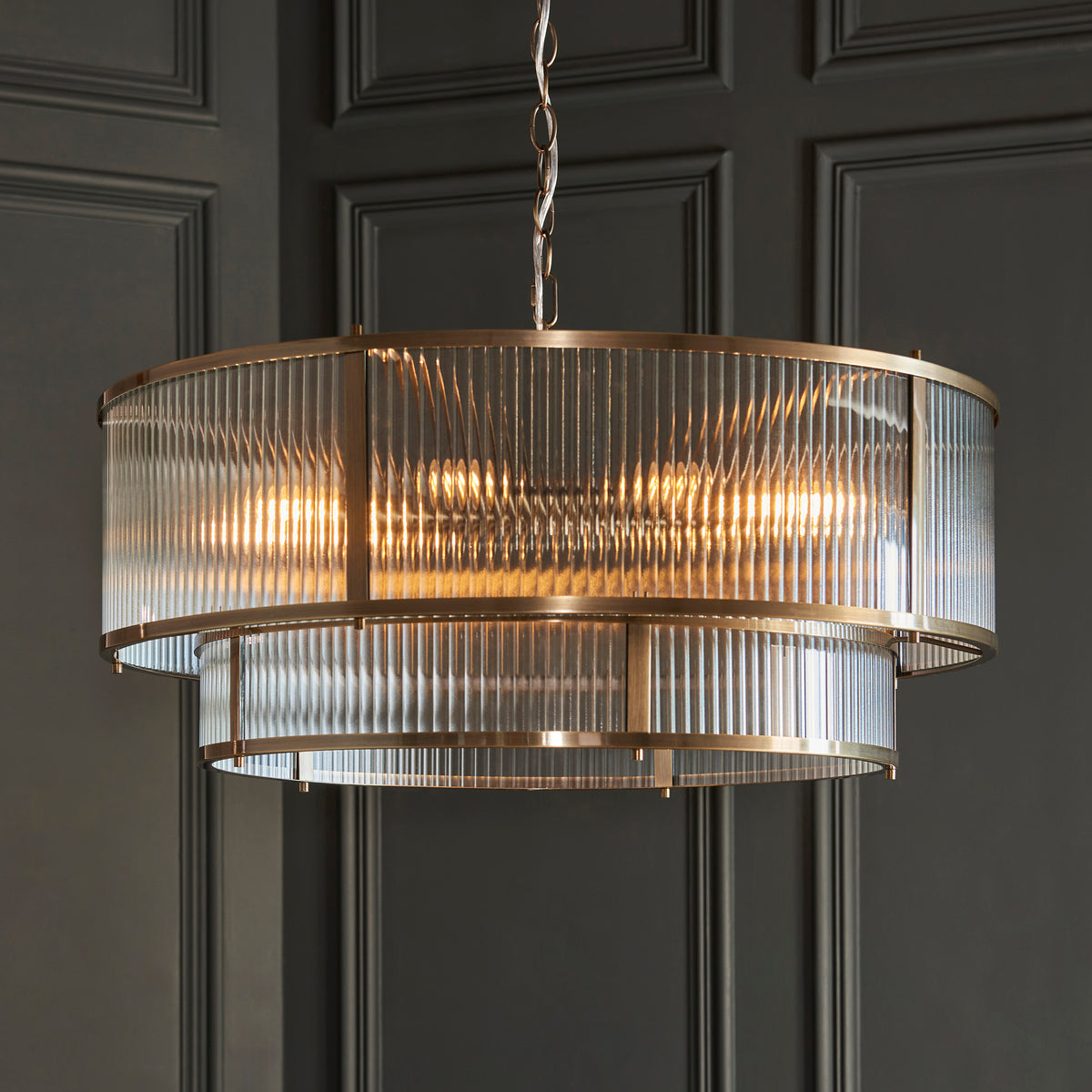 A two-tiered chandelier with ribbed glass and a brass frame hangs against a dark paneled wall.