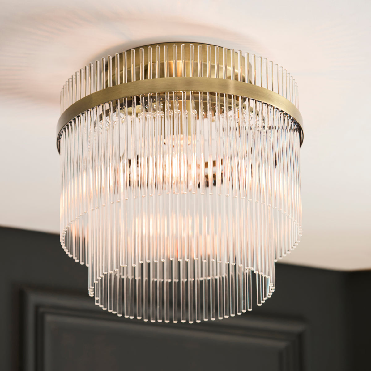 A close-up of a modern ceiling light fixture with a brass frame and clear glass rods, emitting a warm glow.