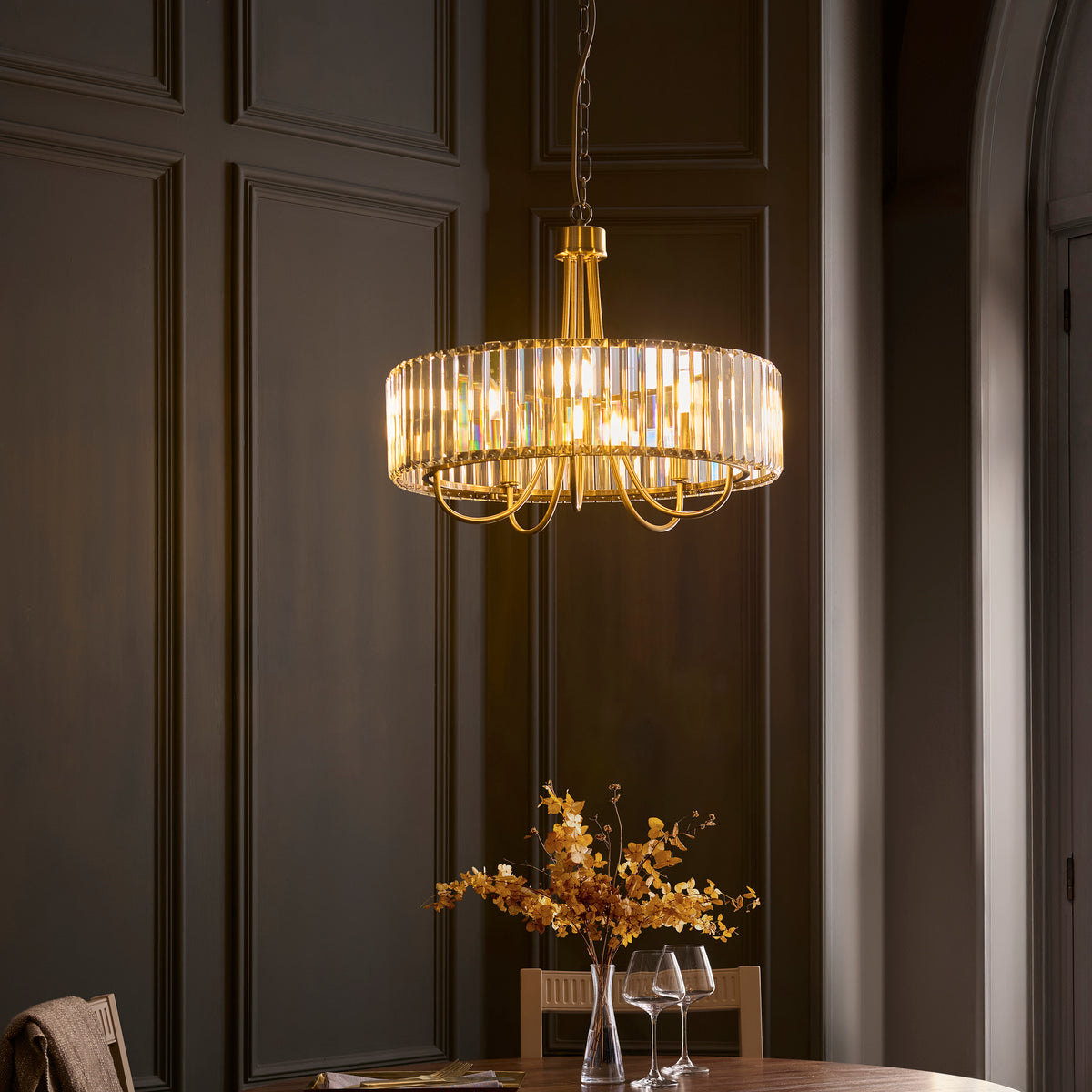 A gold and crystal chandelier hangs above a dining table with a vase of dried flowers and wine glasses.