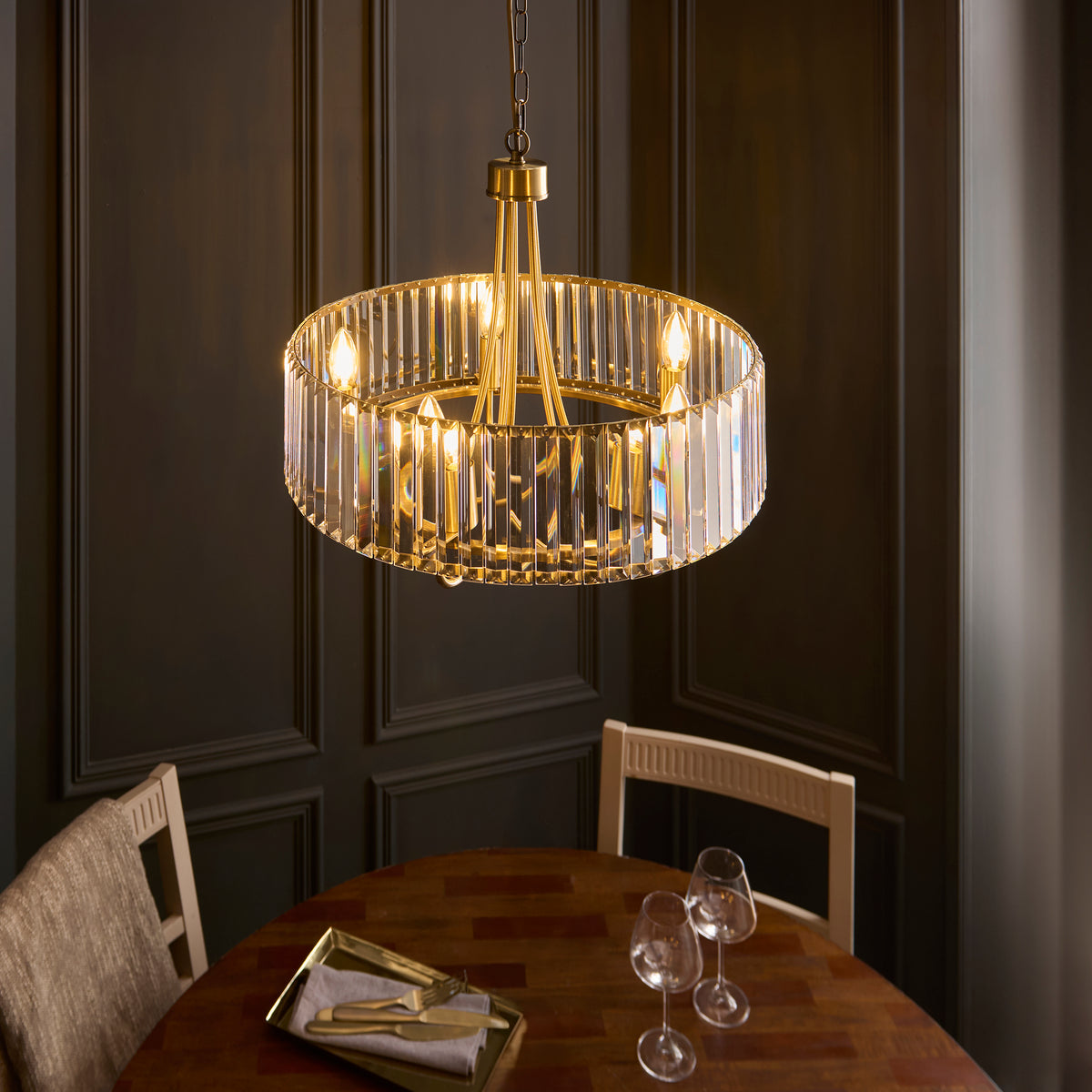 A gold and crystal chandelier hangs above a wooden dining table set with two wine glasses and a tray of gold cutlery.
