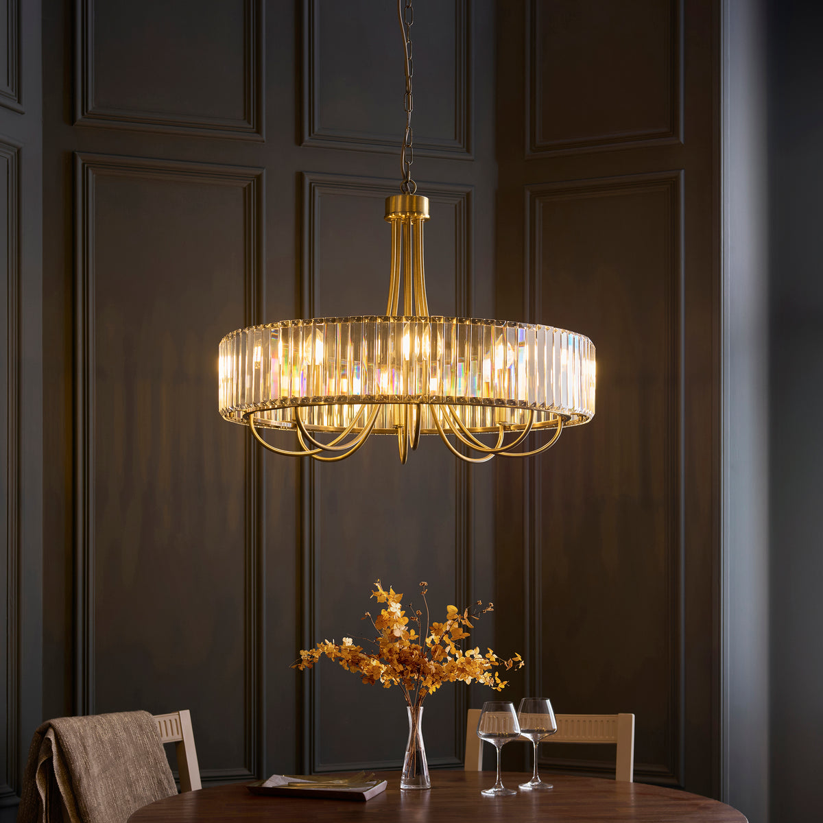 A gold chandelier with a crystal-encrusted drum shade hangs above a dining table with a vase of dried flowers.