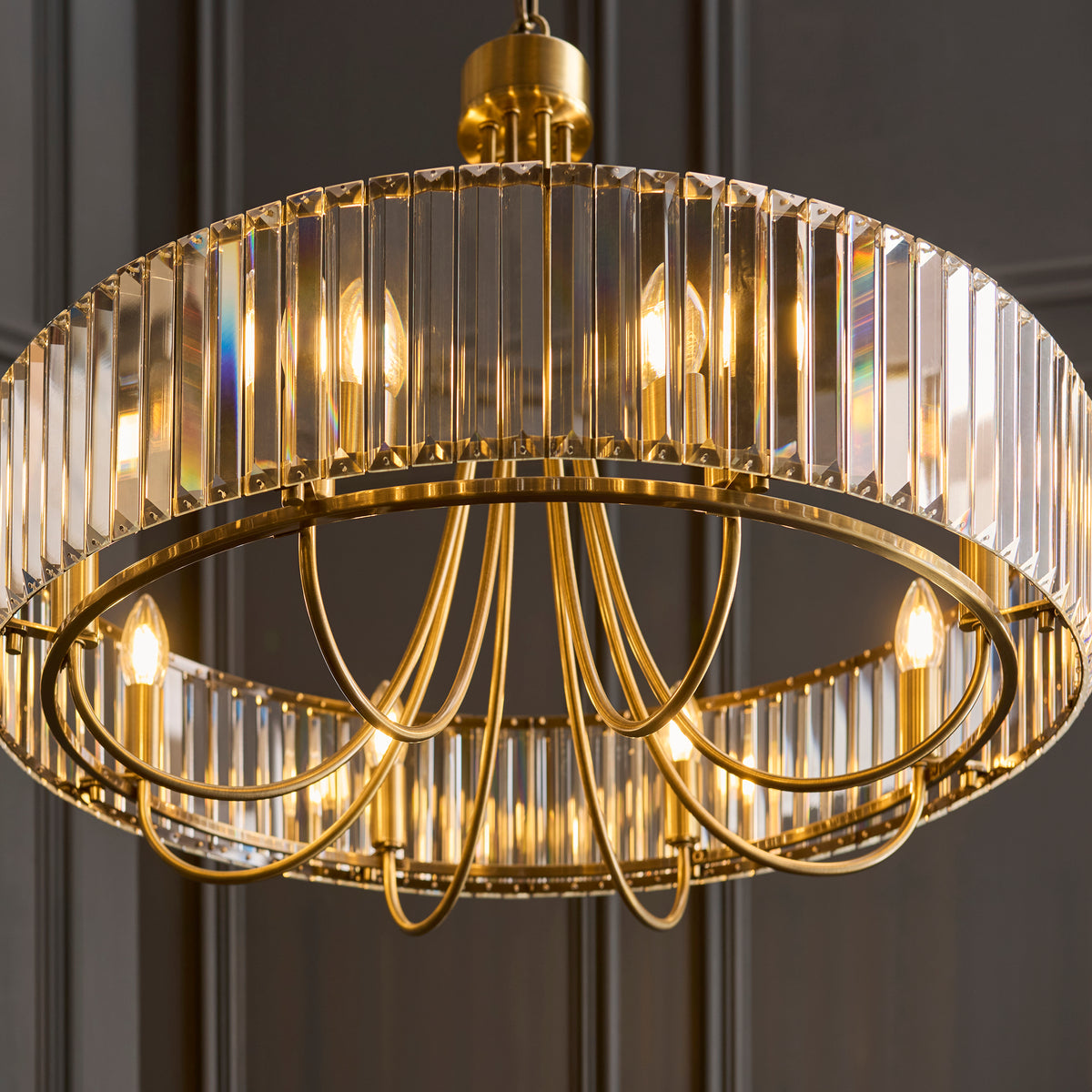 Round brass chandelier with vertical crystal panels and exposed bulbs, against a dark paneled wall. 