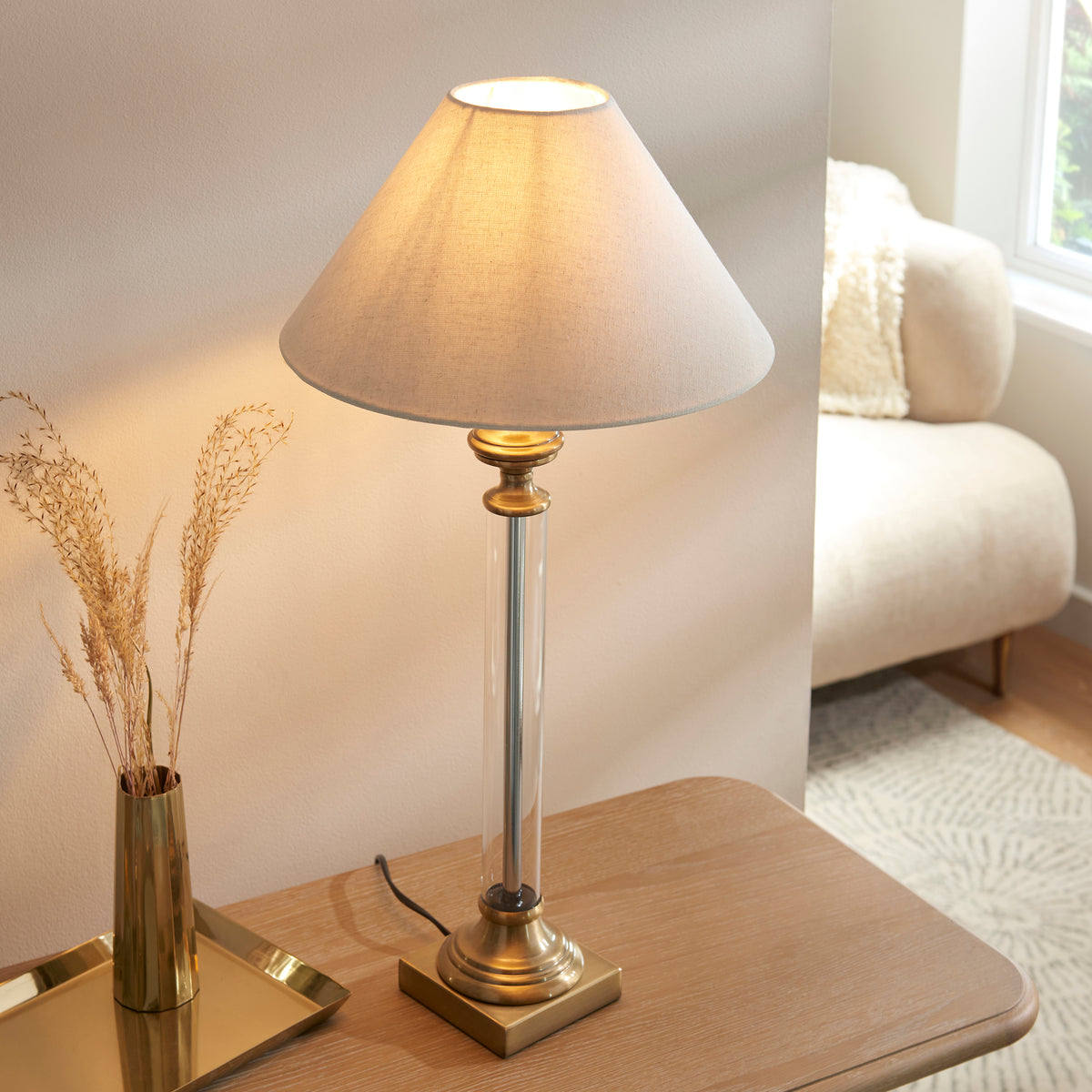 A brass and clear glass table lamp with a light fabric shade, next to a gold vase with dried grasses.