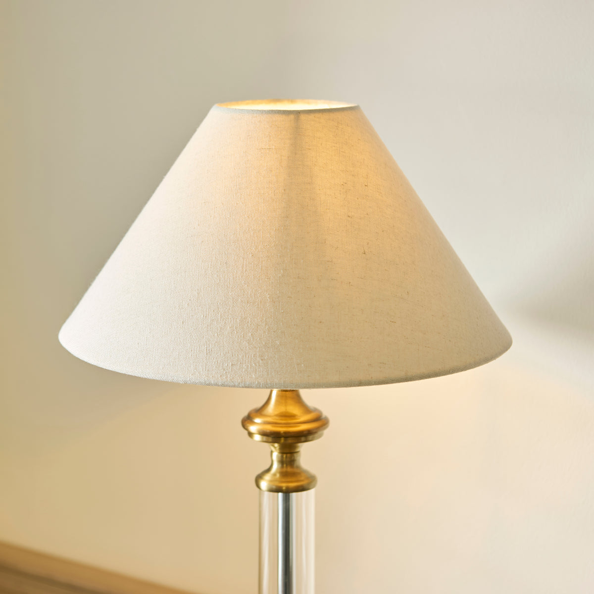 A close-up of a table lamp with a beige fabric shade and a brass and clear glass base.