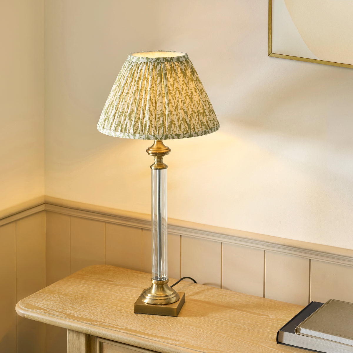 A table lamp with a pleated green and white patterned shade, clear glass column, and brass base on a wooden table.
