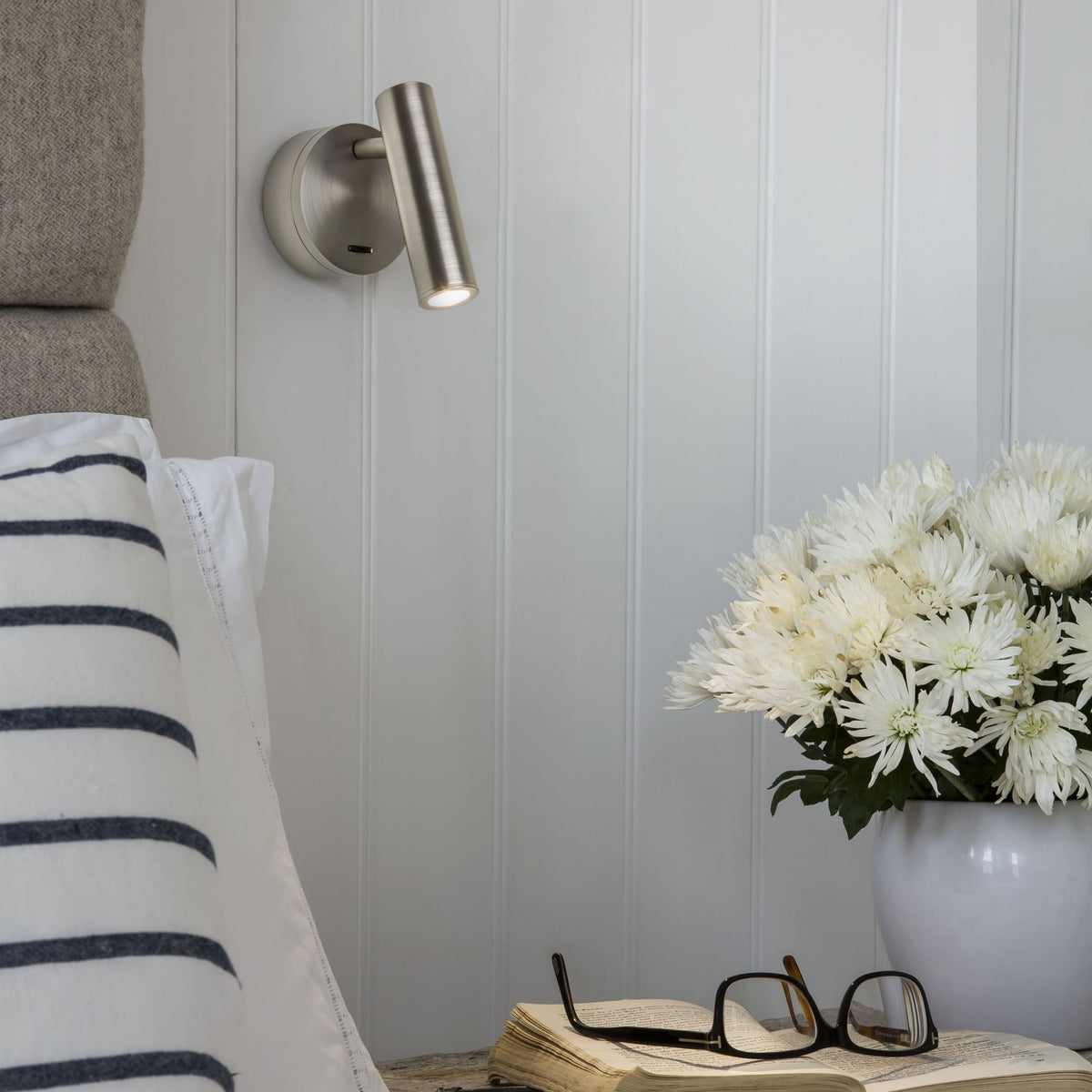 Brushed steel wall light with a lit bulb, above an open book, reading glasses, and white flowers