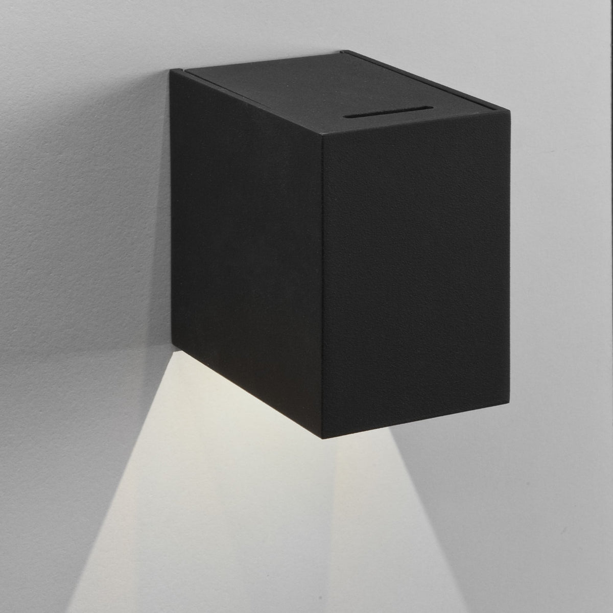 Black rectangular wall light with a slot on top, casting a downward beam of light on a white wall