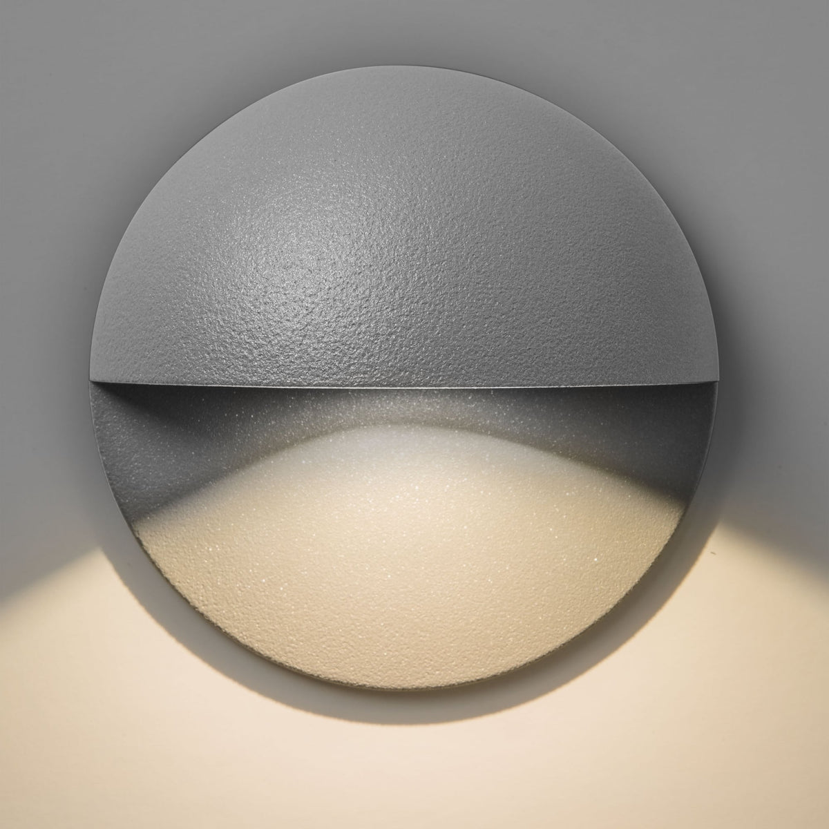 Round grey wall light with a textured finish, emitting a warm glow downwards onto a light surface