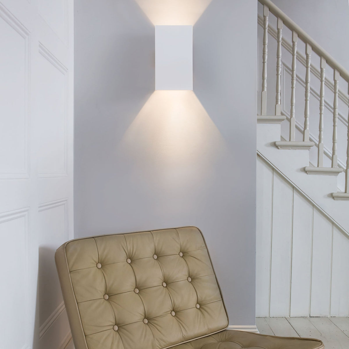 White rectangular wall light illuminating a light grey wall above a beige tufted leather chair