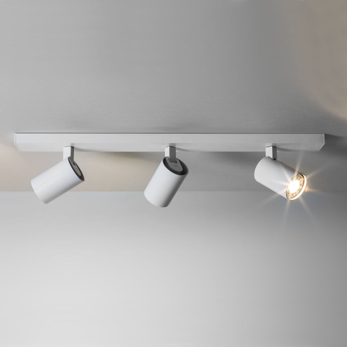 Three white cylindrical track lights on a white ceiling track, one light is illuminated