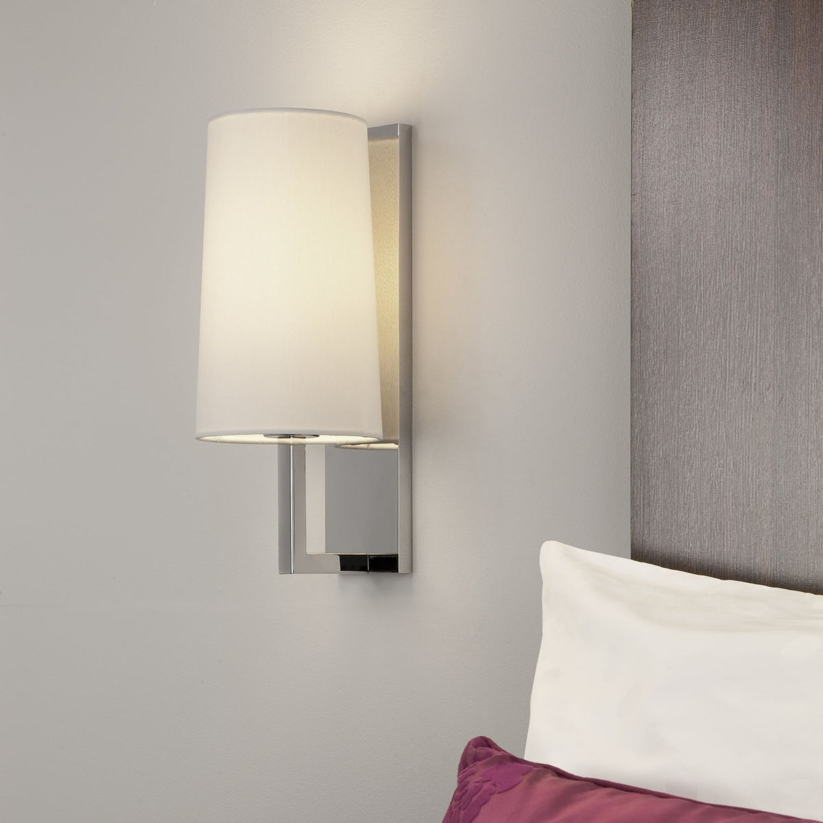 Wall-mounted lamp with a white shade and a chrome base, next to a bed with a white pillow and a purple cushion