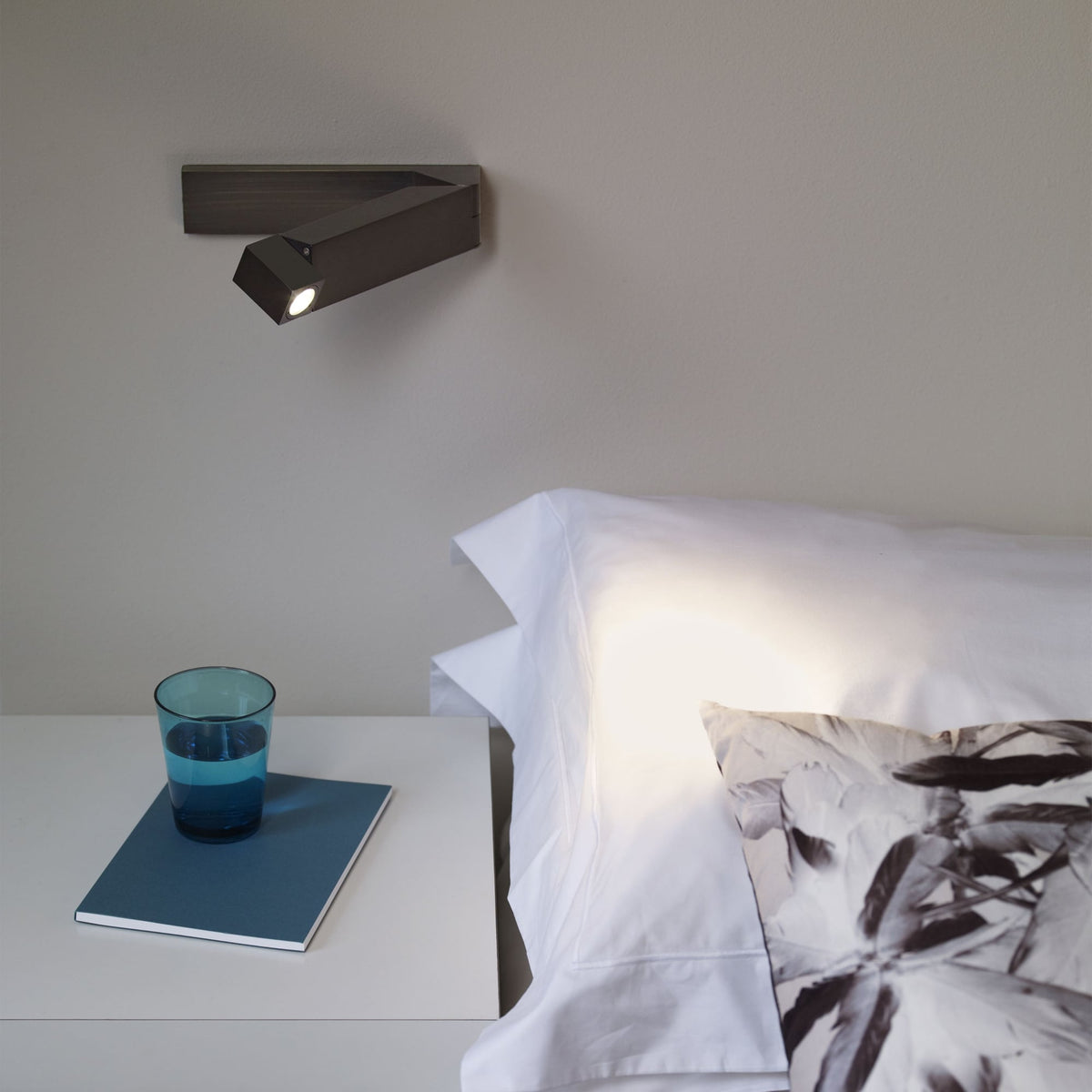 Wall-mounted reading light above a bed with white sheets, a patterned pillow, and a glass of water on a nightstand