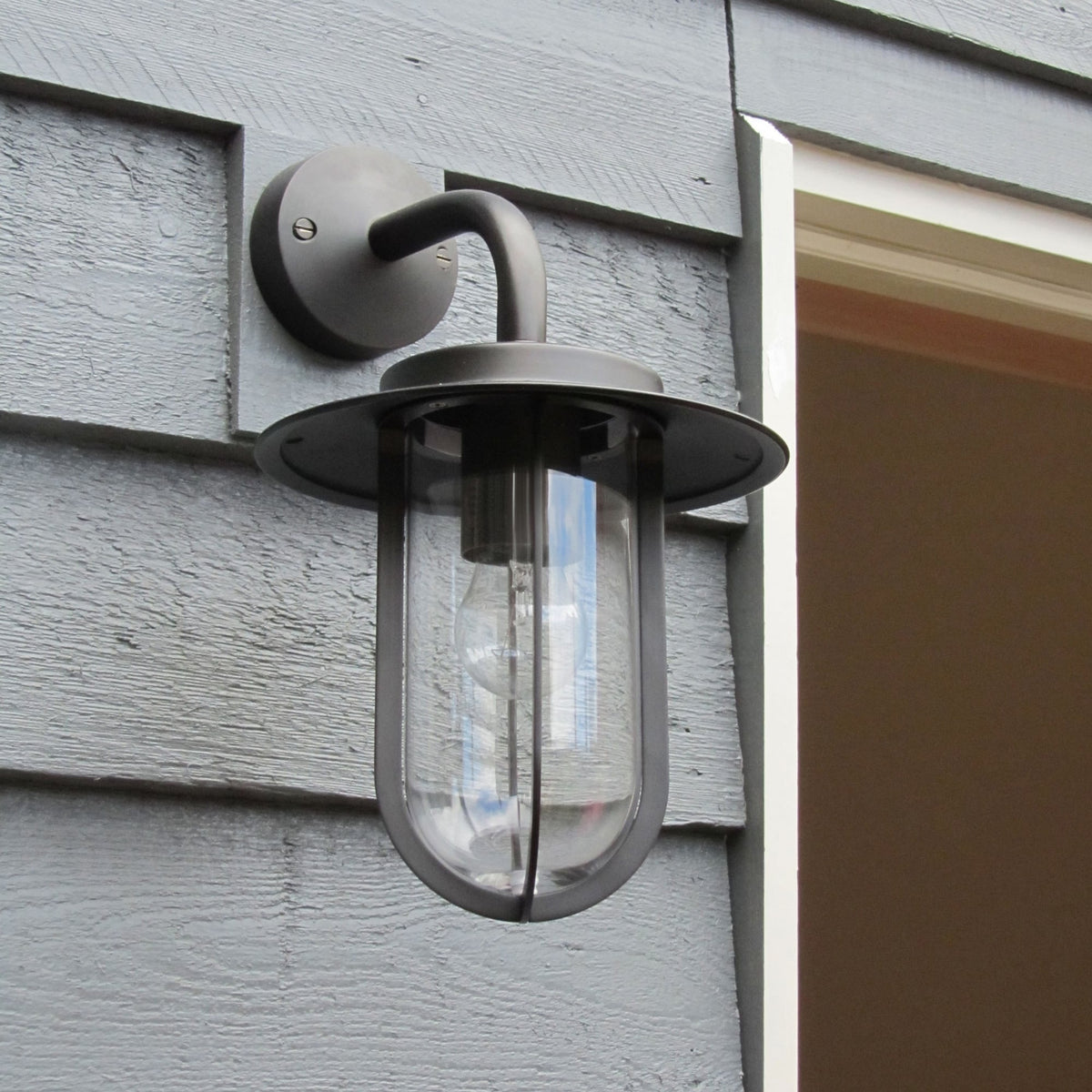Outdoor wall light with a dark bronze finish and clear glass, mounted on gray siding.