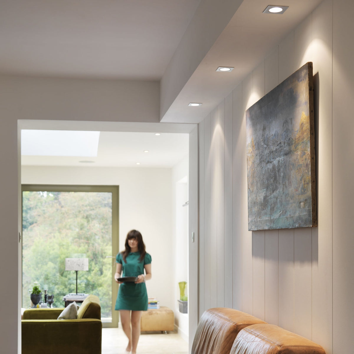 Woman in green dress walks through a modern living room with recessed lighting and a large painting