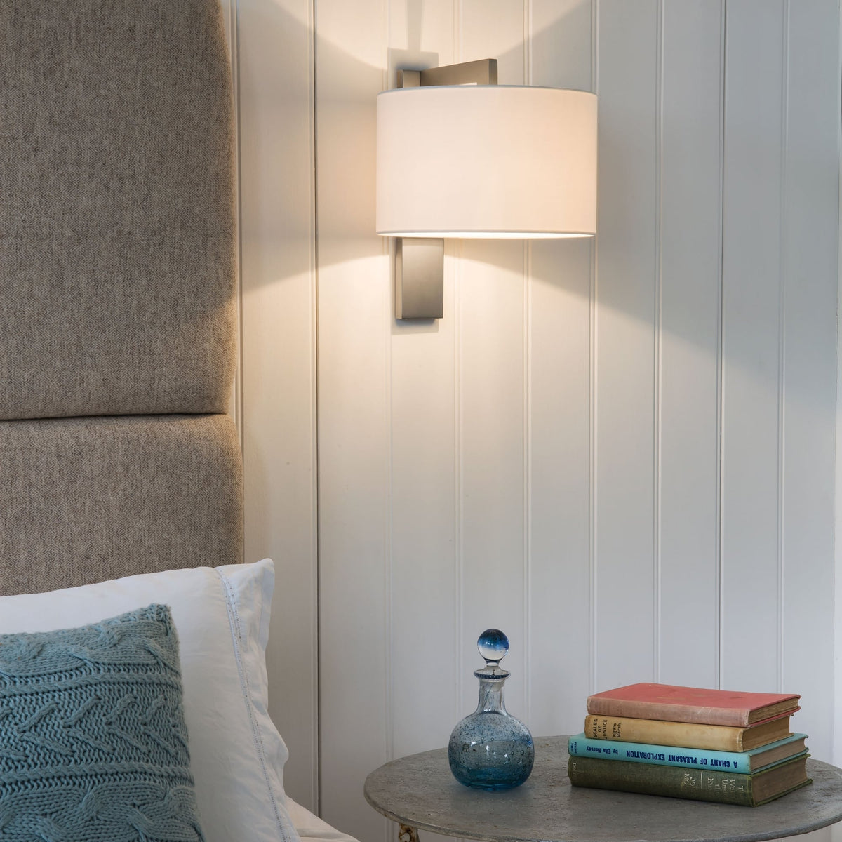 Wall light with a white shade and a grey base, above a bedside table with books and a blue glass bottle