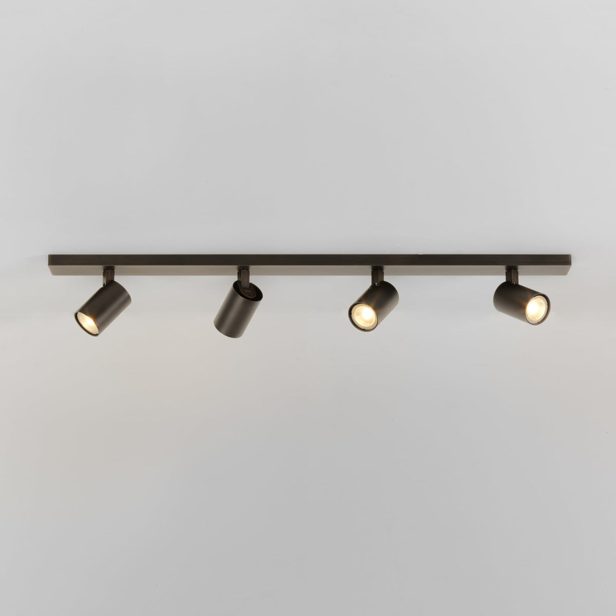Four dark bronze cylindrical track lights on a matching track bar, emitting warm light against a white ceiling