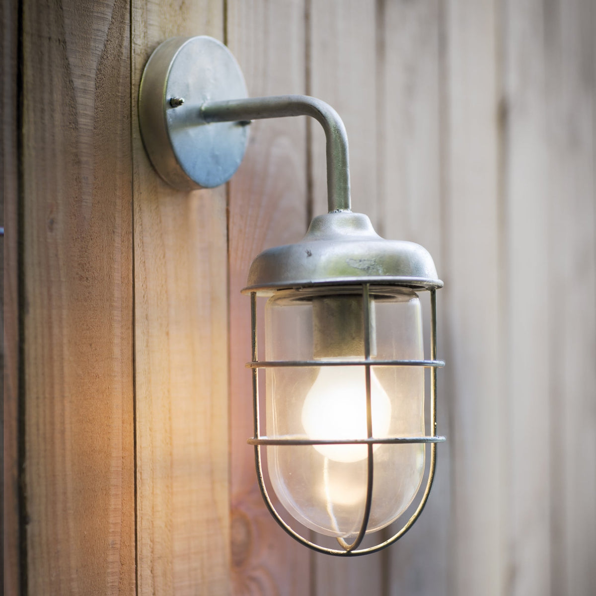 A caged outdoor wall light with an exposed bulb, fixed to a wooden fence.