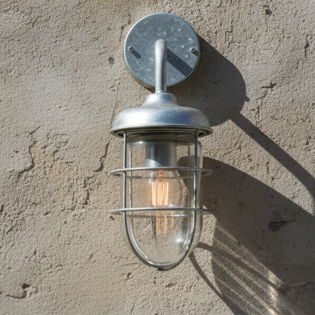 Galvanized outdoor wall light with a clear glass shade and exposed filament bulb on a textured wall.
