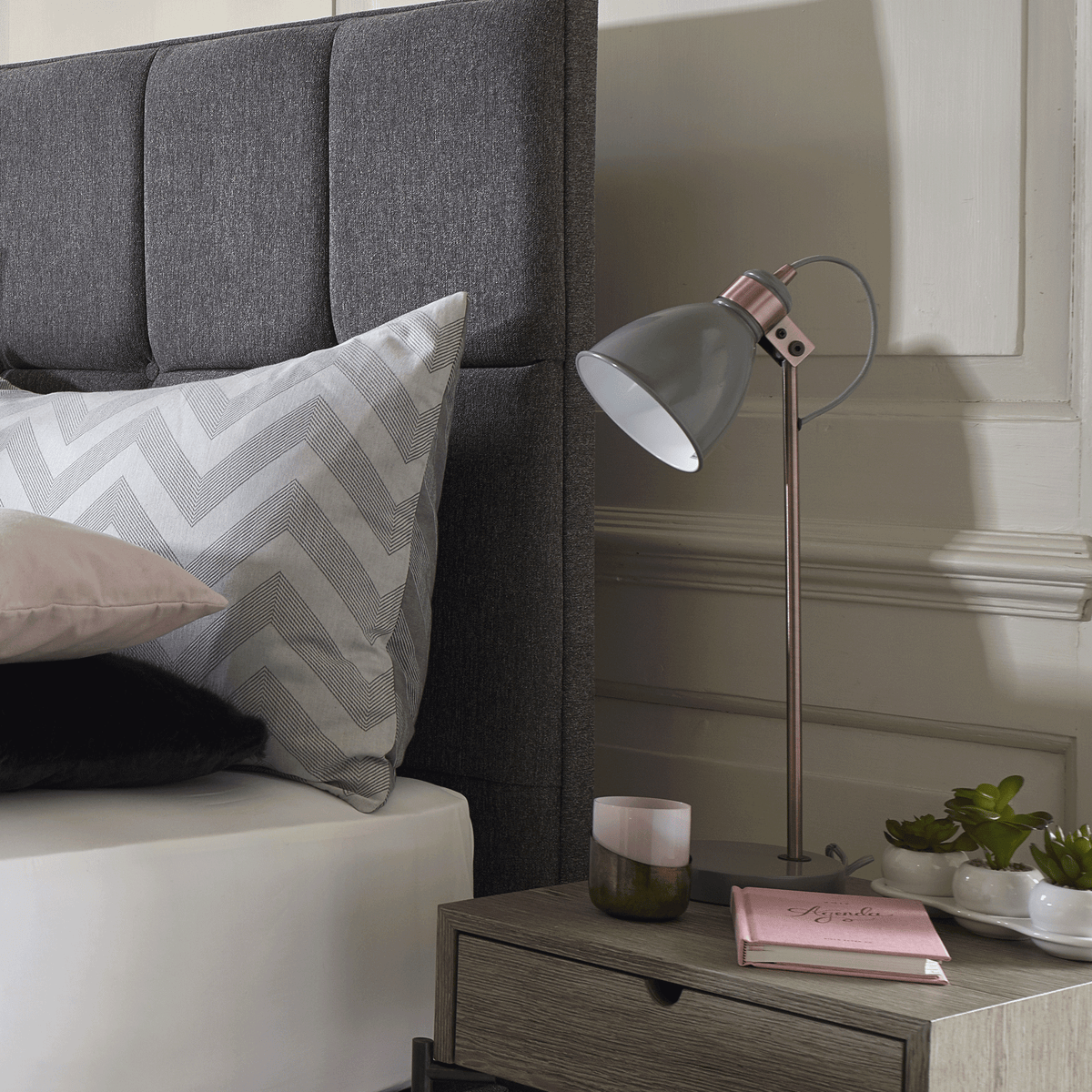 A gray upholstered headboard with pillows, a nightstand with a lamp, and decorative items.
