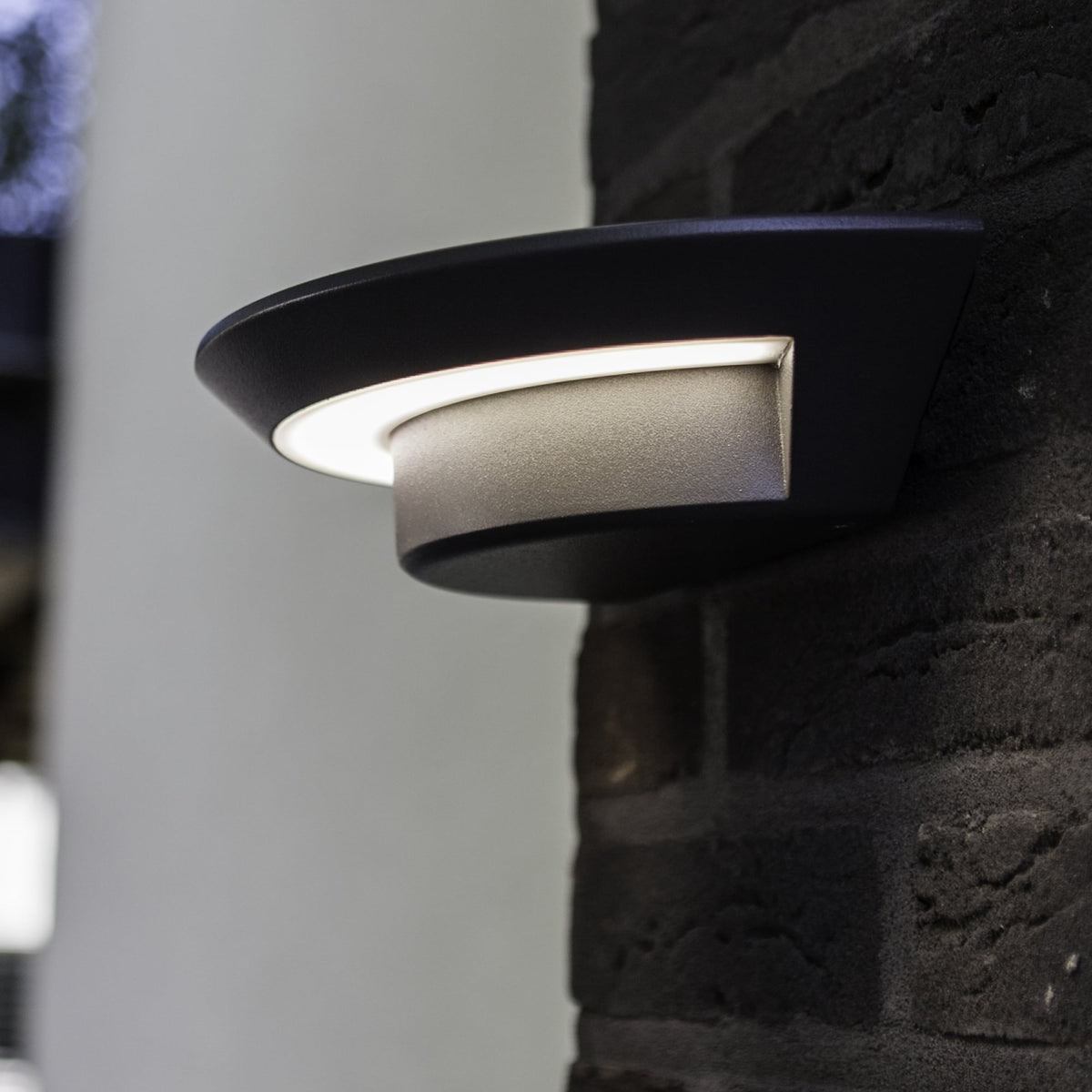Modern black outdoor wall light with a bright, curved LED strip, mounted on a dark brick wall.