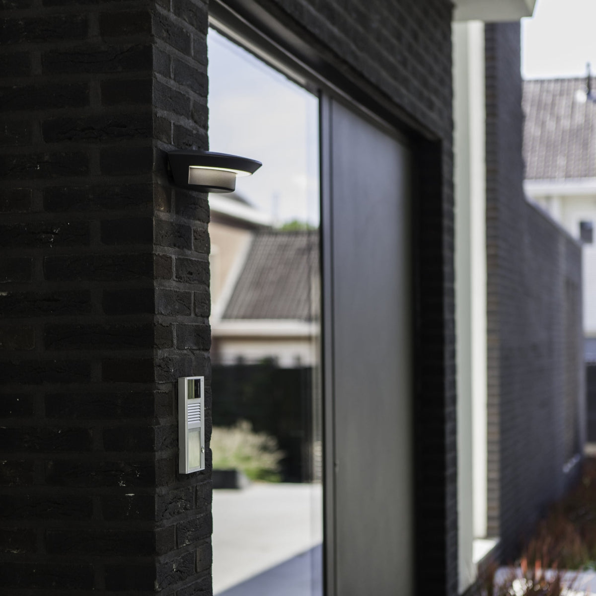 Modern outdoor light and intercom system on a dark brick wall next to a large window.